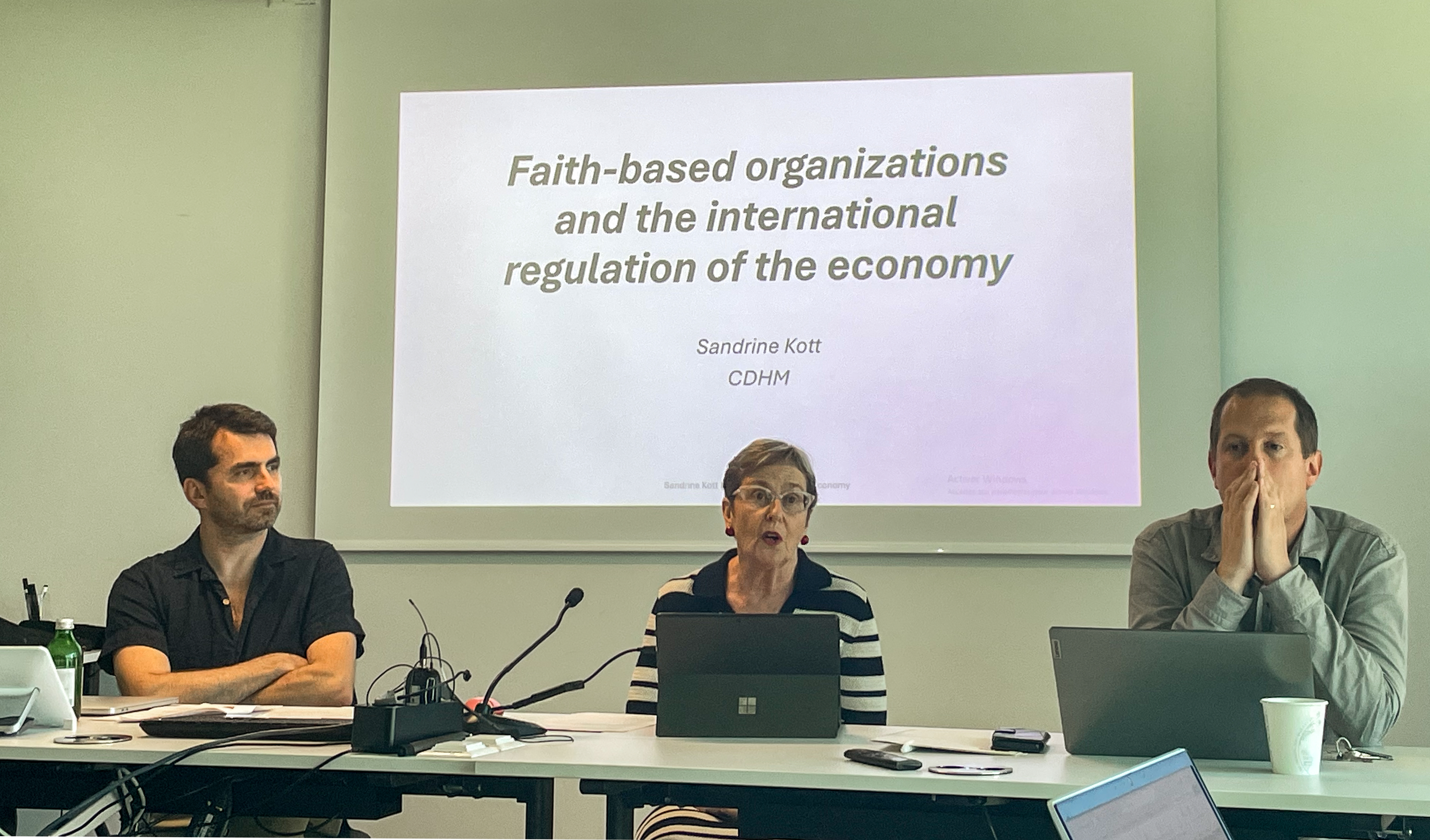 Three people seated at a conference table, with a presentation slide titled "Faith-based organizations and the international regulation of the economy" projected behind. The atmosphere is professional.