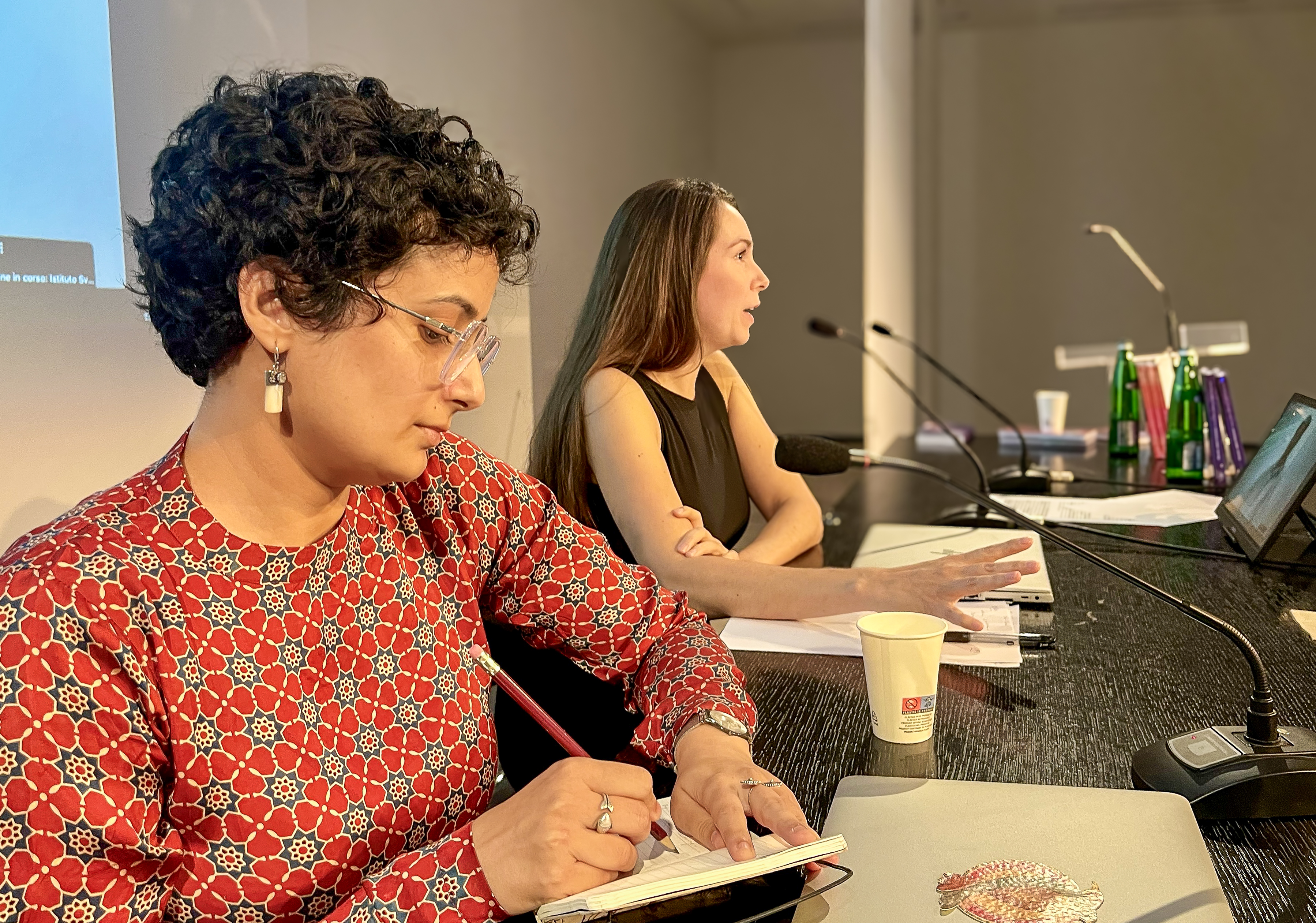 Two women at a conference sit at a table with microphones. One writes in a notebook, while the other speaks. The setting appears focused and professional.