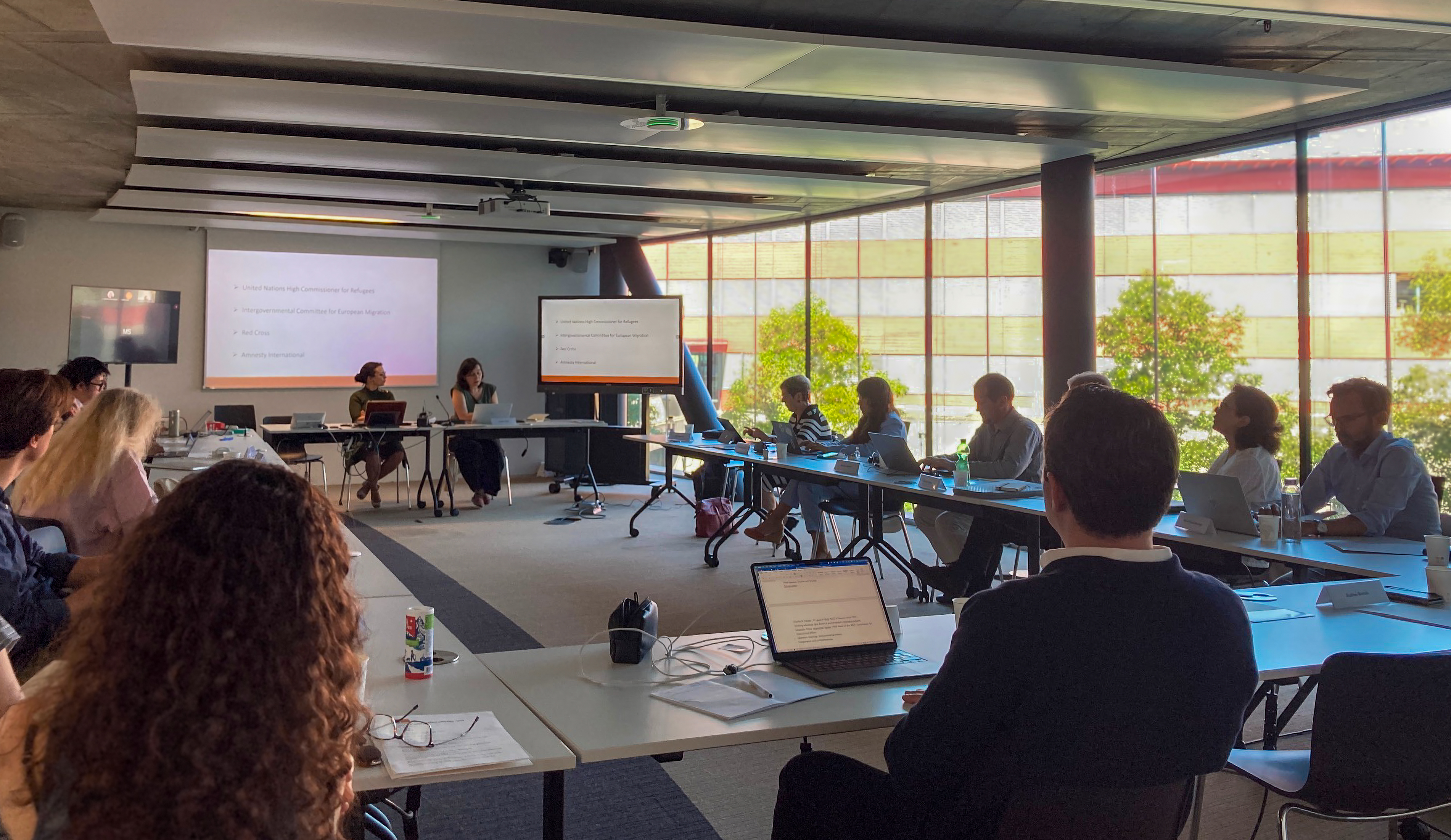 A group of people are seated around a U-shaped conference table in a modern, well-lit room, attentively listening to a presentation.