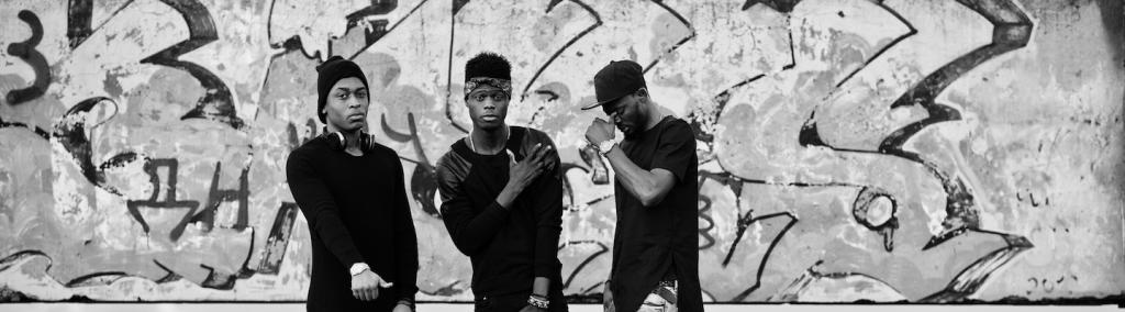 Three rap singers on a roof; background graffiti wall.