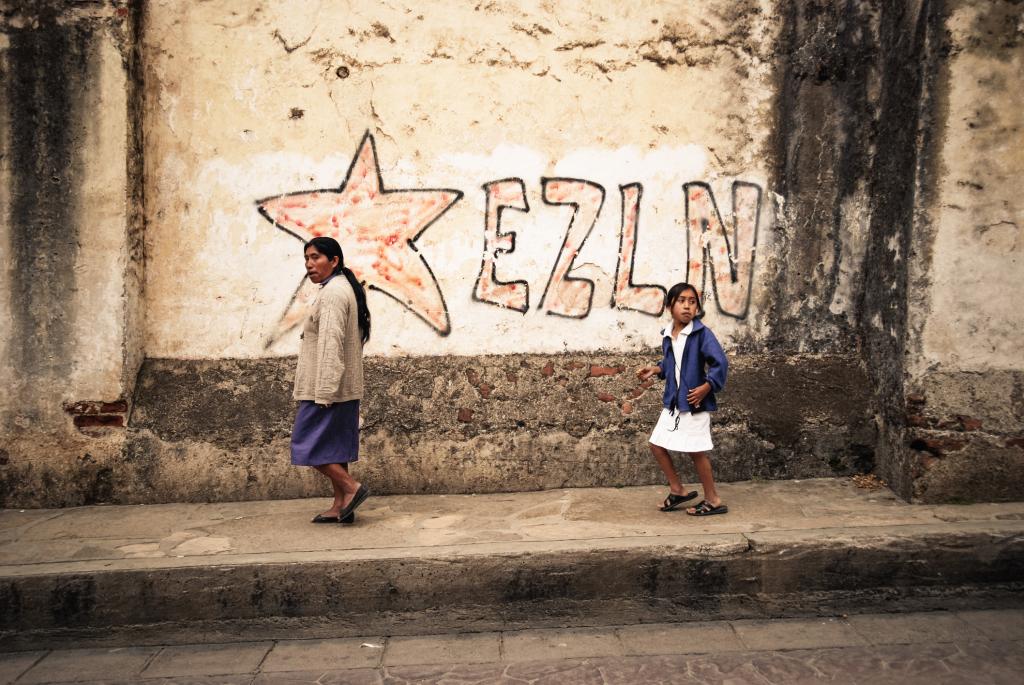 Two Mayans walk next to a wall with the inscription EZLN (Ej!AA>rcito Zapatista de Liberacion Nacional, EZLN), Zapatista Army of National Liberation."