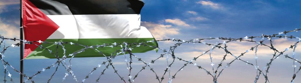 A fence with barbed wire against a colourful sky and a Palestinian flagpole.