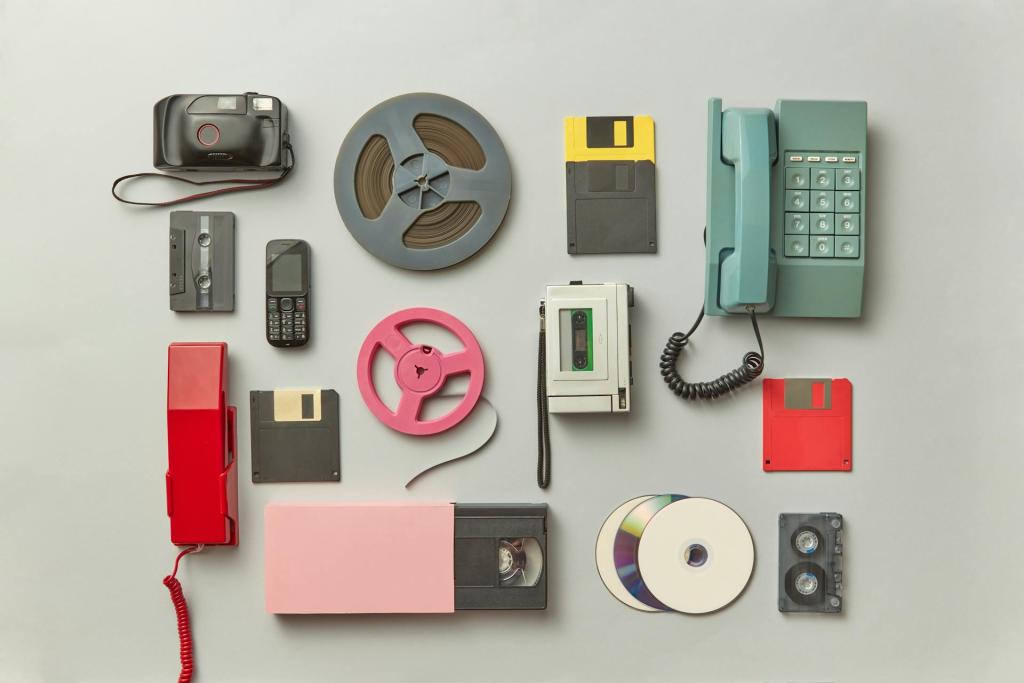 Vintage technology items arranged on a light surface, including a film camera, floppy disks, rotary phone, pager, CD, cassette, and an analog phone.