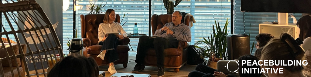 A man and woman sit in armchairs, engaged in discussion, surrounded by listeners in a modern office. The logo reads "Peacebuilding Initiative."