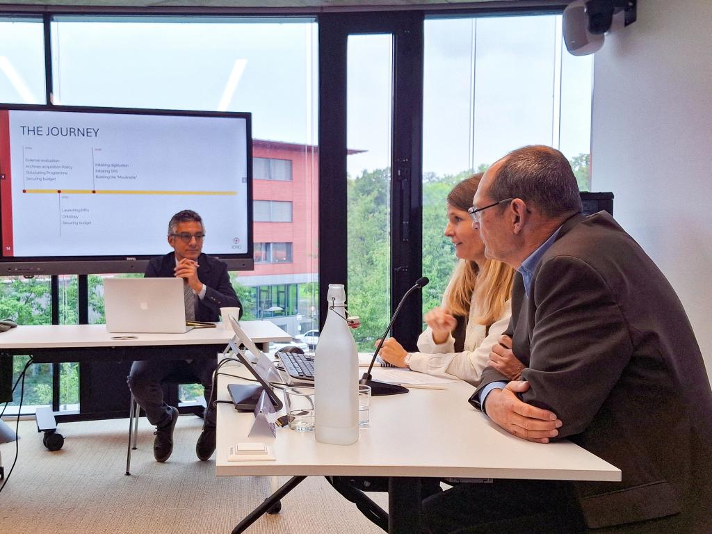 Three people in a meeting room discussing near a large screen displaying a presentation titled "The Journey." The setting is professional and focused.