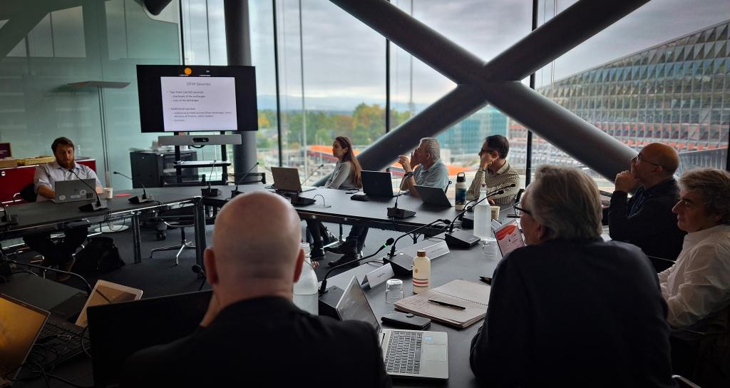 A group of people sit around a large conference table in a modern office, attentively watching a presentation on a screen. The room has large windows offering a view of a cloudy sky and a geometric building. The atmosphere appears focused and collaborative.