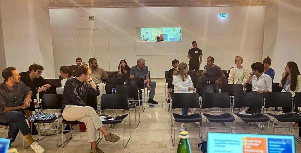 A group of people seated in rows of black chairs in a bright room, engaged in conversation. A presentation screen is visible in the foreground.
