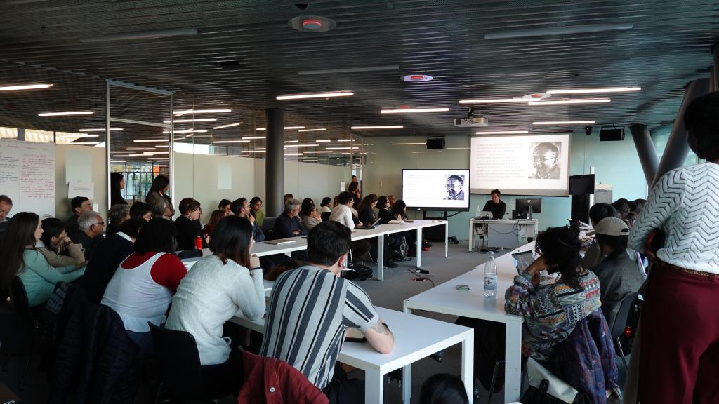 A diverse group of people sits around white tables in a modern conference room, attentively listening to a speaker at the front with a presentation screen.