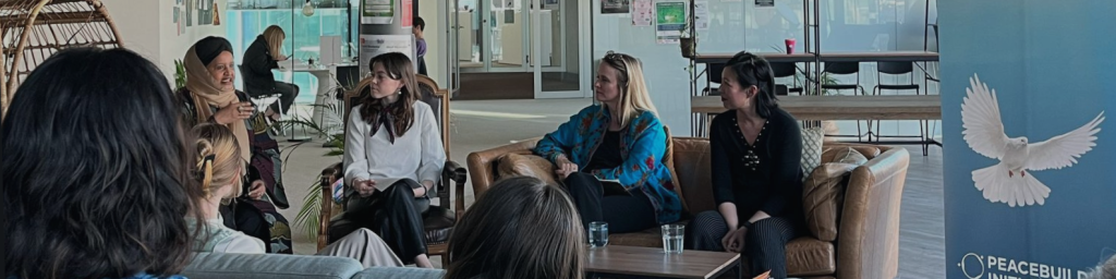 Four women sit on couches in a modern, open space, engaged in discussion. A "Peacebuilding Initiative" banner with a dove is visible. The atmosphere is collaborative.