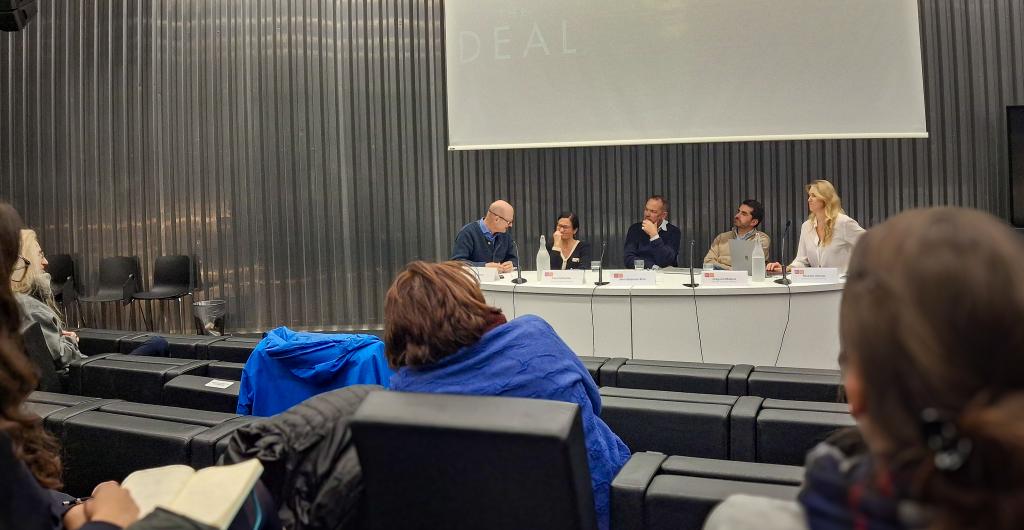 A panel of five people sits at a long table in a modern lecture hall. Audience members watch and take notes, creating an engaging and focused atmosphere.