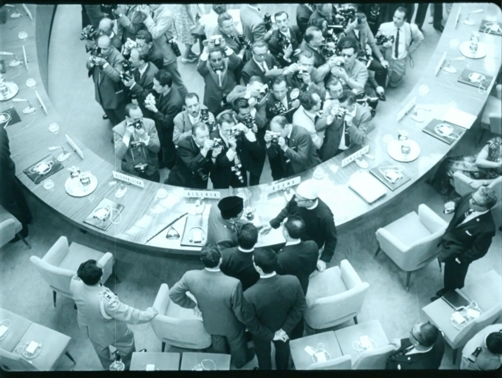 Overhead view of a crowded conference room, photographers surround delegates shaking hands across a circular table. 