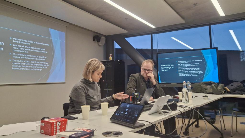 A woman and a man discuss at a conference table with laptops, papers, and cups. A presentation titled "Humanitarian Knowledge in Danger" is on screens. The atmosphere is focused and professional.
