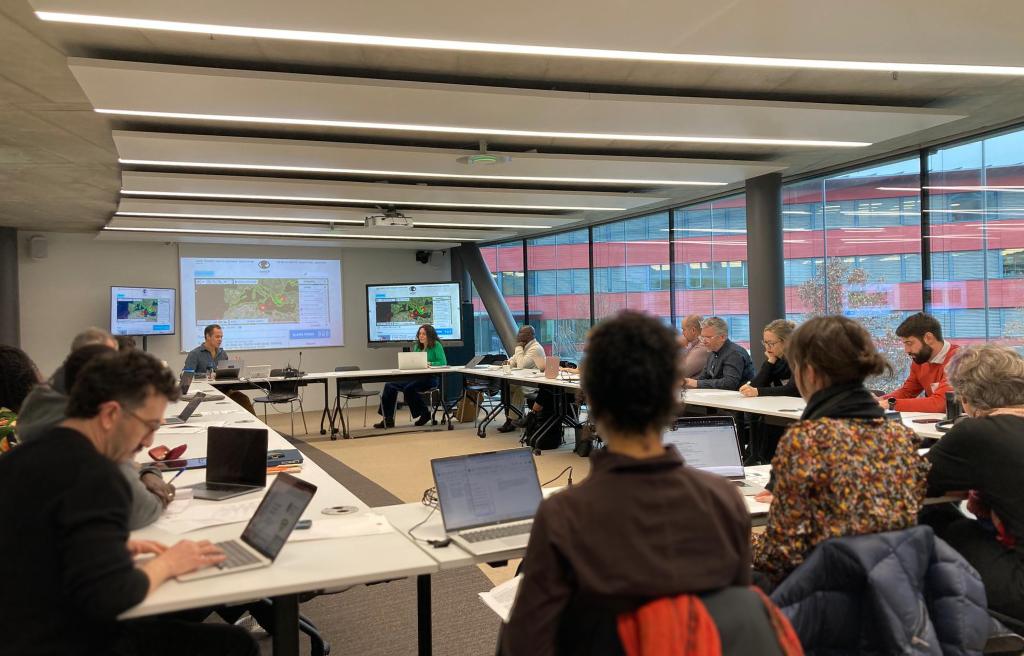 A group of people sit around a U-shaped table in a modern conference room. They are focused on a presentation displayed on two screens.