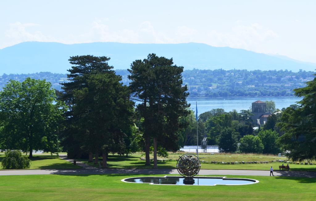 View of Ariana park with lush green trees and a reflective pond featuring the celestial sphere. A lake and distant mountains under a clear sky form the backdrop.
