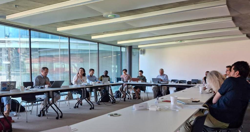 A group of people sit at a long U-shaped table in a modern conference room with large windows. The atmosphere appears professional and collaborative.