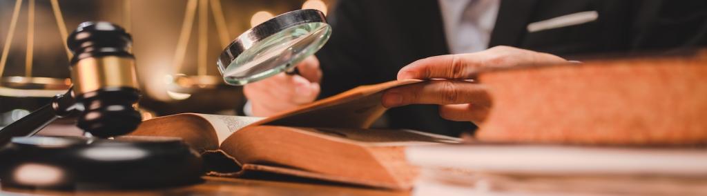 A judge’s gavel sits on a table beside an open book, over which a hand is holding a magnifying glass.