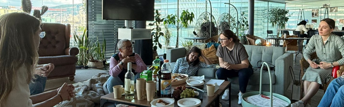 A group of people gathered in the fab. They're seated around a coffee table with drinks and snacks, engaged in conversation. The atmosphere is relaxed and friendly.