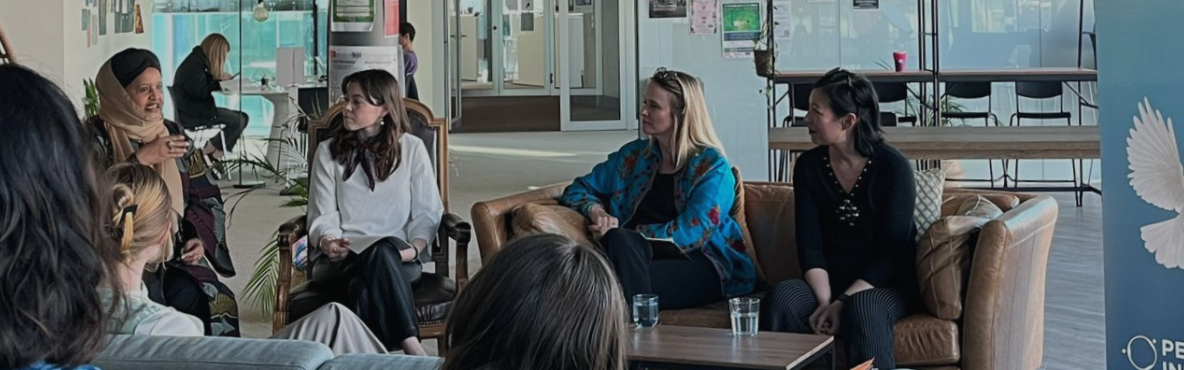 Four women sit on couches in a modern, open space, engaged in discussion. A "Peacebuilding Initiative" banner with a dove is visible. The atmosphere is collaborative.
