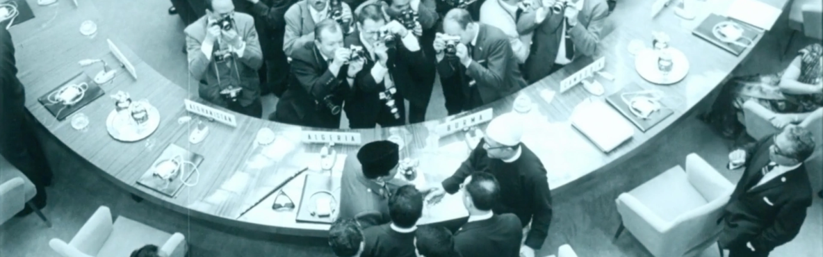 Overhead view of a crowded conference room, photographers surround delegates shaking hands across a circular table. 