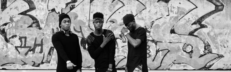 Three rap singers on a roof; background graffiti wall.