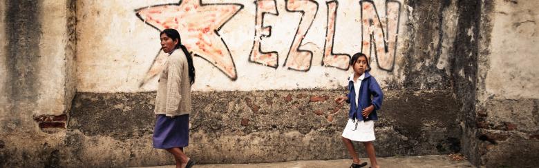 Two Mayans walk next to a wall with the inscription EZLN (Ej!AA>rcito Zapatista de Liberacion Nacional, EZLN), Zapatista Army of National Liberation."