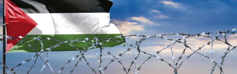 A fence with barbed wire against a colourful sky and a Palestinian flagpole.