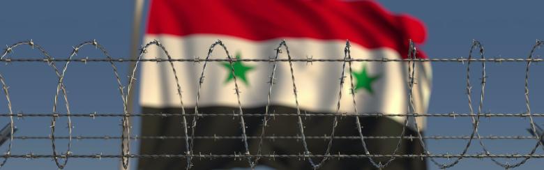 Blurred waving flag of Syria behind barbed wire fence.