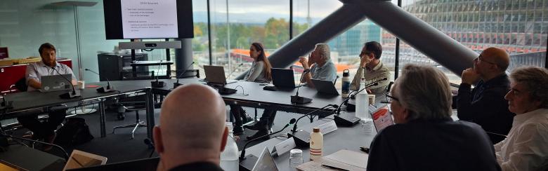A group of people sit around a large conference table in a modern office, attentively watching a presentation on a screen. The room has large windows offering a view of a cloudy sky and a geometric building. The atmosphere appears focused and collaborative.
