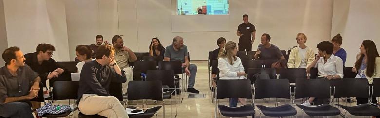 A group of people seated in rows of black chairs in a bright room, engaged in conversation. A presentation screen is visible in the foreground.