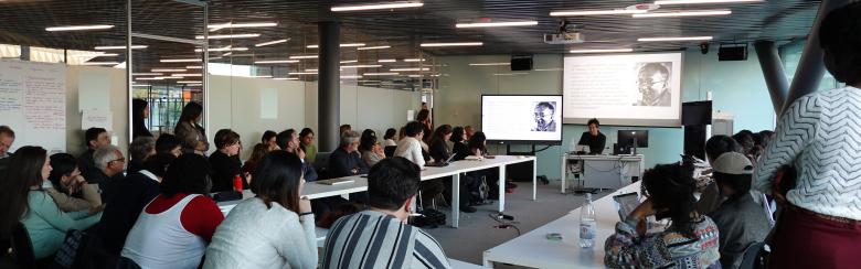 A diverse group of people sits around white tables in a modern conference room, attentively listening to a speaker at the front with a presentation screen.