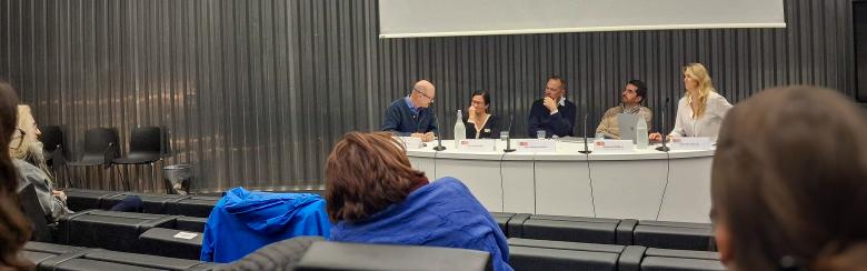 A panel of five people sits at a long table in a modern lecture hall. Audience members watch and take notes, creating an engaging and focused atmosphere.