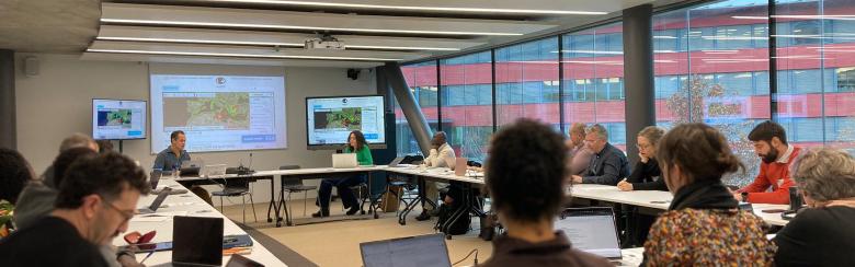 A group of people sit around a U-shaped table in a modern conference room. They are focused on a presentation displayed on two screens.