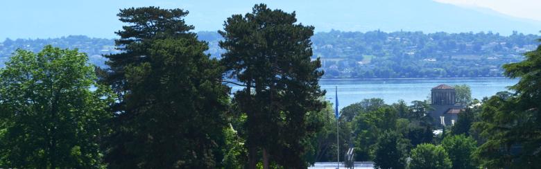 View of Ariana park with lush green trees and a reflective pond featuring the celestial sphere. A lake and distant mountains under a clear sky form the backdrop.