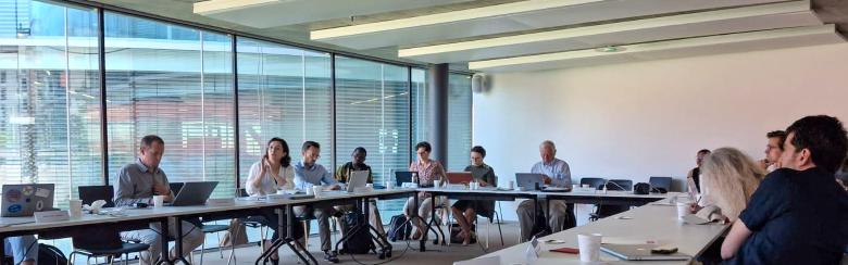 A group of people sit at a long U-shaped table in a modern conference room with large windows. The atmosphere appears professional and collaborative.