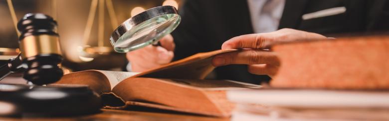 A judge’s gavel sits on a table beside an open book, over which a hand is holding a magnifying glass.